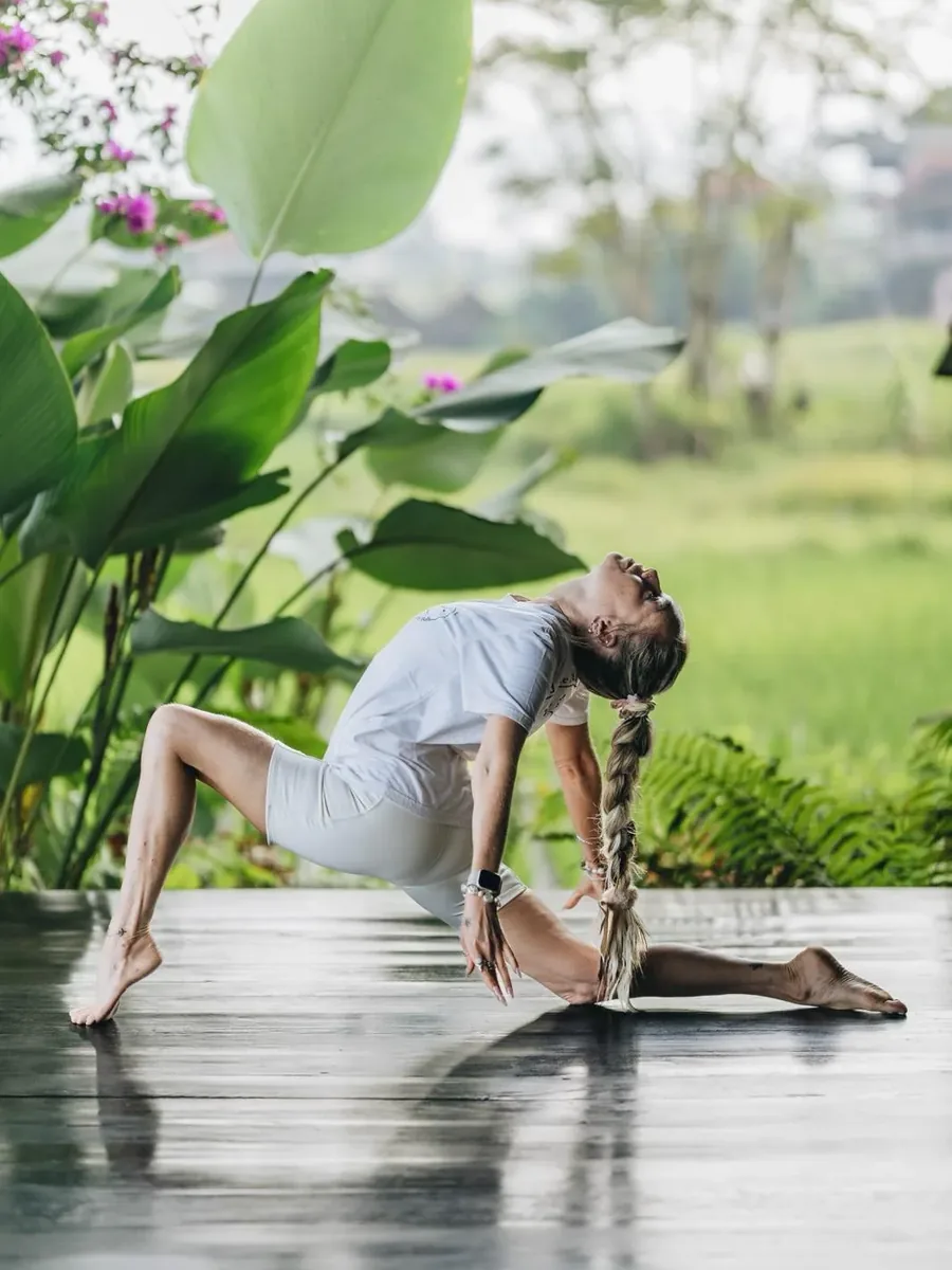 A woman in a gentle backbend on a rain-wet wooden deck beside rice fields in Bali