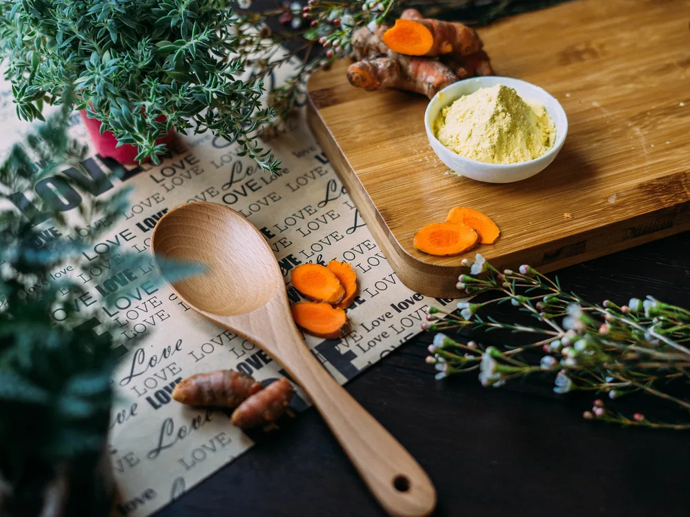 Fresh turmeric slices, a wooden spoon, herbs and a bowl of spice powder on a chopping board